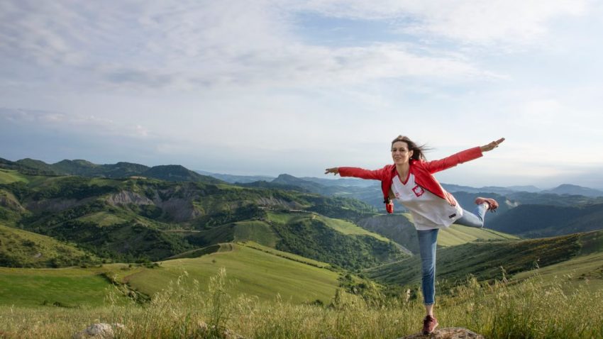 A woman joyfully balances on a rock amidst rolling green hills under a clear blue sky.
