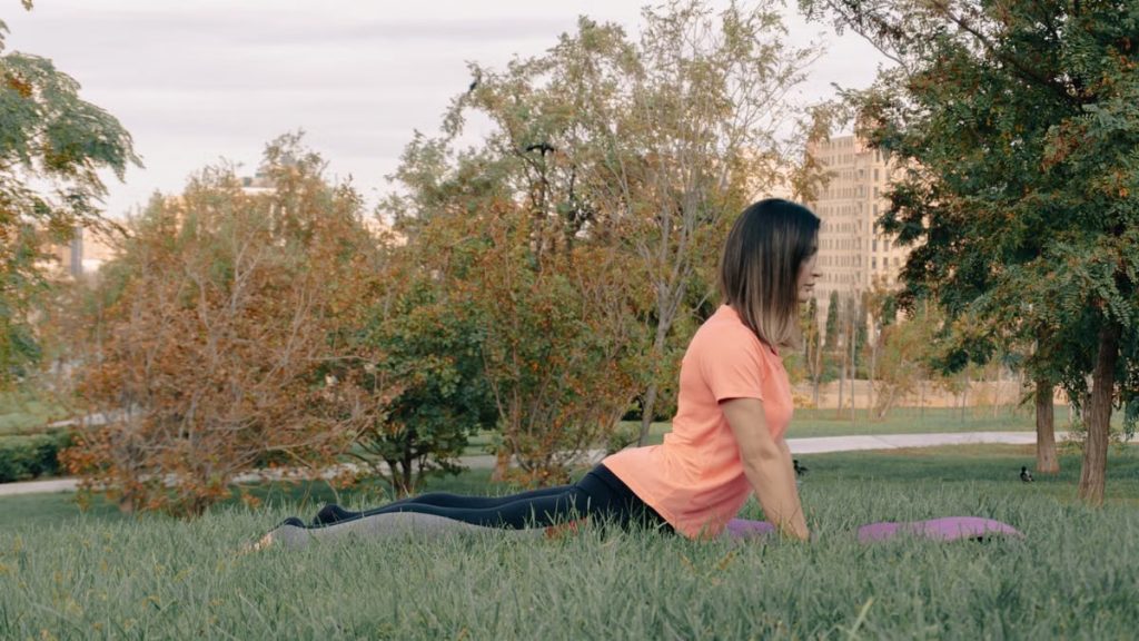 Adult woman performing yoga exercise outdoors in a peaceful park setting, surrounded by greenery.