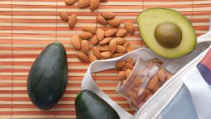 Top view of fresh avocados and almonds in a glass jar on a bamboo mat, perfect for healthy snacking.